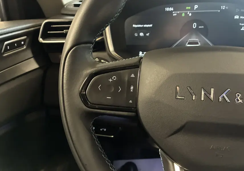 Vue rapprochée du volant noir de la LYNK & CO 01 avec commandes intégrées et tableau de bord digital en arrière-plan.