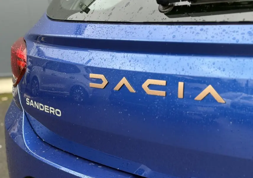 Gros plan arrière gauche sur le coffre bleu Iron de la Dacia Sandero avec lettrage cuivré sous la pluie.