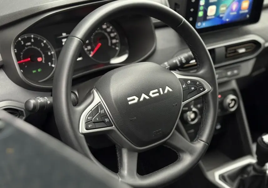 Vue rapprochée du volant noir Dacia avec commandes intégrées et écran tactile du tableau de bord en arrière-plan.