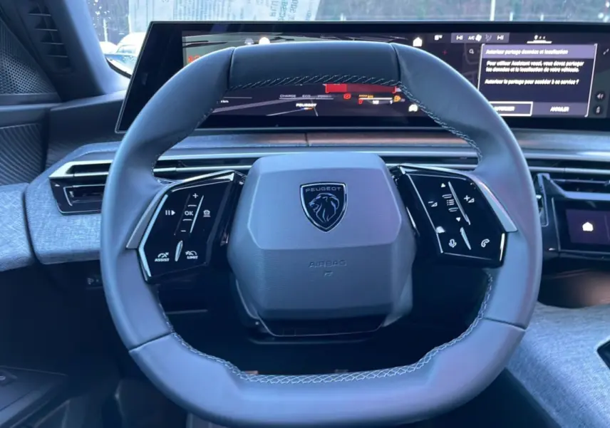 Vue rapprochée du volant cuir multifonctions gris avec logo Peugeot, écran numérique panoramique en arrière-plan.