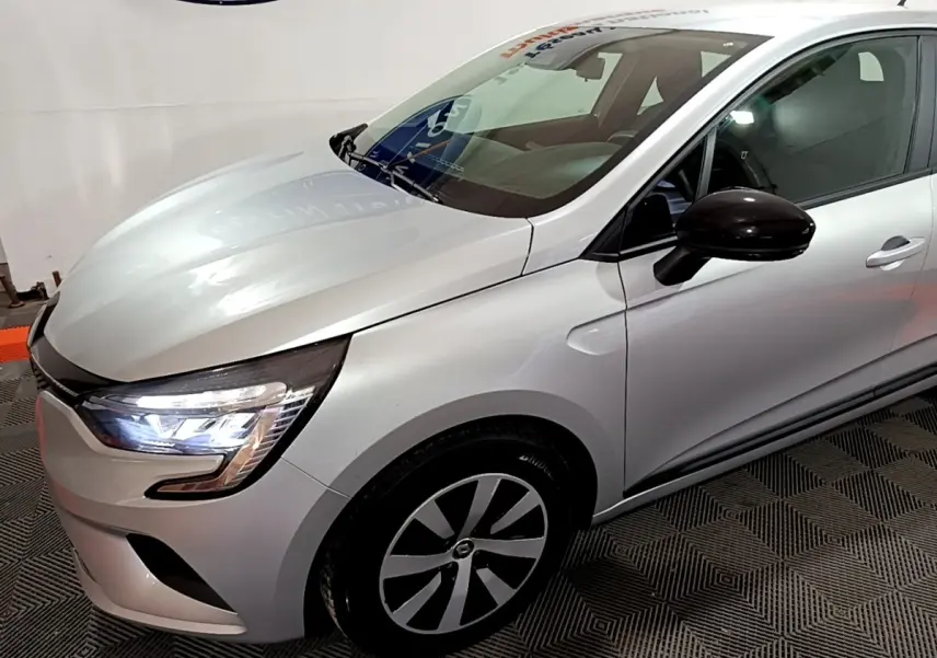 Vue 3/4 avant droit d'une Renault Clio gris clair 2023 avec phares LED allumés et rétroviseur noir brillant.