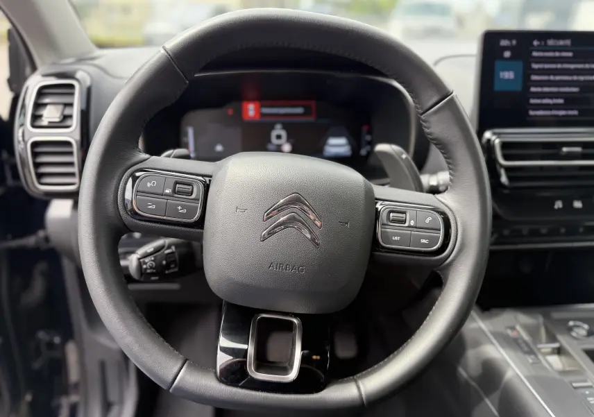 Gros plan sur le volant noir du Citroën C5 Aircross Hybrid 2025 avec commandes intégrées et tableau de bord numérique flou.
