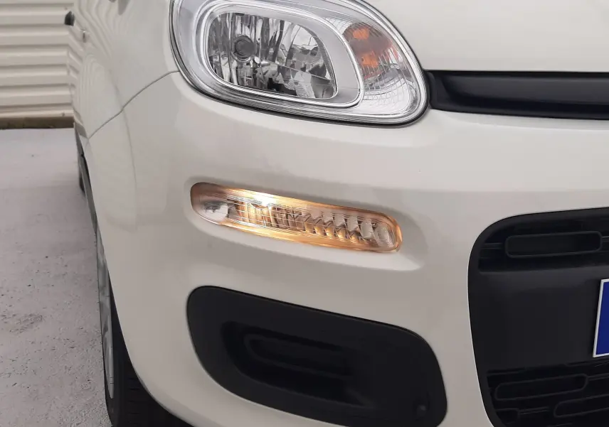 Gros plan sur le phare avant gauche et le clignotant allumé d'une Fiat Panda 2025 blanc gelato, avec pare-chocs noir.