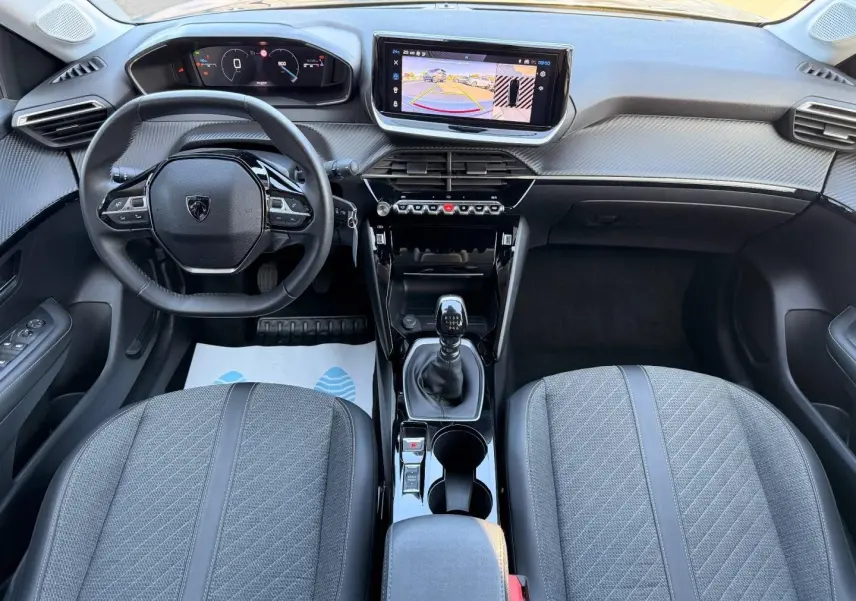 Vue intérieure avant de la Peugeot 208 1.2 essence 2025, volant compact, écran tactile et sièges tissu gris.