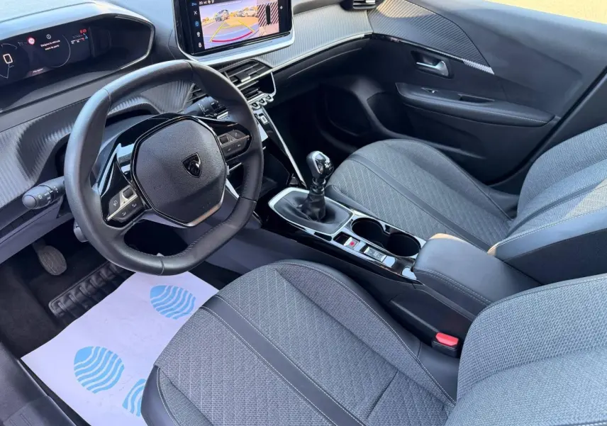 Vue intérieure côté conducteur de la Peugeot 208 1.2 essence 2025, volant compact et sièges tissu gris avec console centrale noire.
