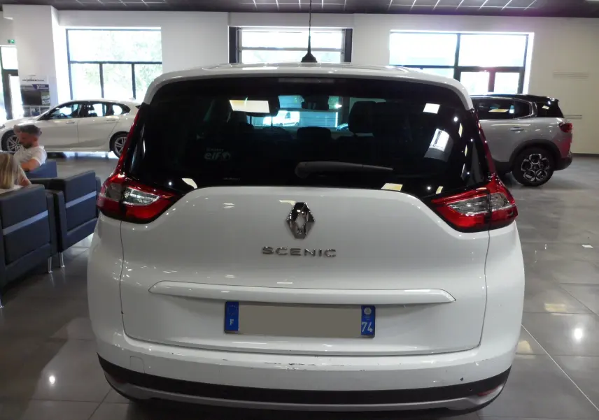 Vue arrière d'un Renault Grand Scenic IV blanc Glacier en intérieur, avec feux arrière rouges et logo Renault visible.