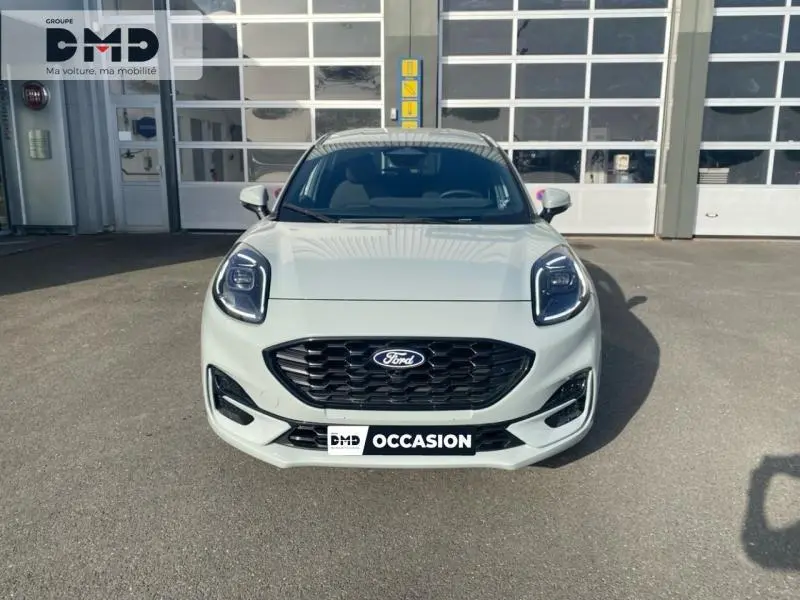 Vue frontale d'une Ford Puma gris clair avec calandre noire et phares LED distinctifs, stationnée devant un garage.