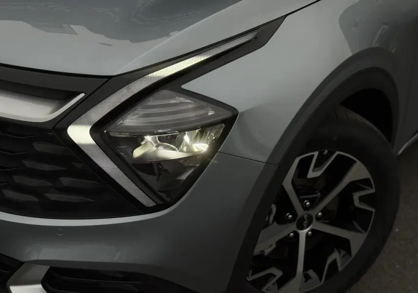 Gros plan sur le phare avant droit allumé et la jante alliage 18'' du Kia Sportage gris perle 2025.