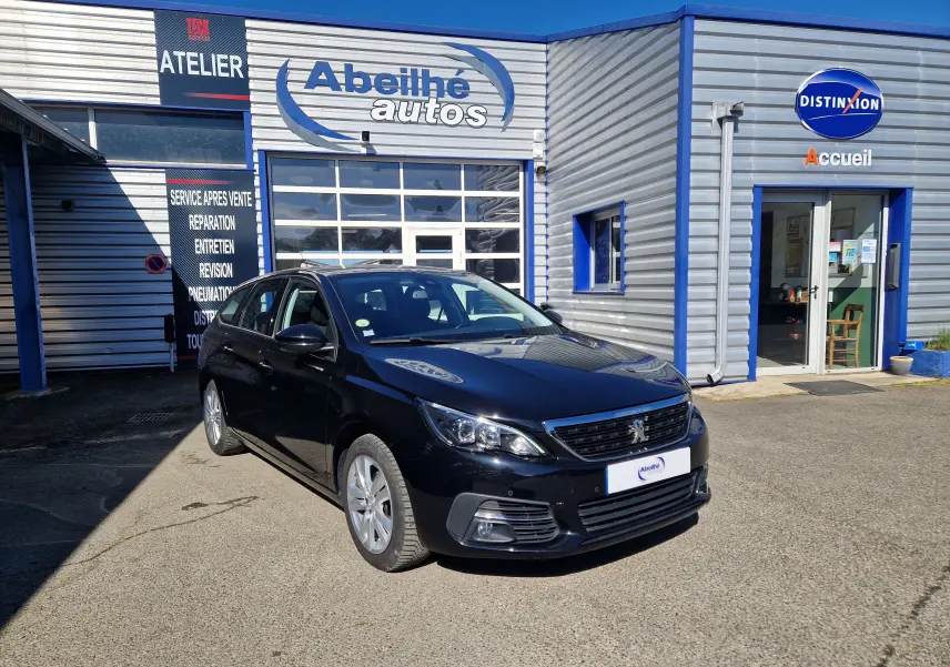 Peugeot 308 SW noire vue en 3/4 avant droit devant un garage avec enseigne Abeilhé autos.