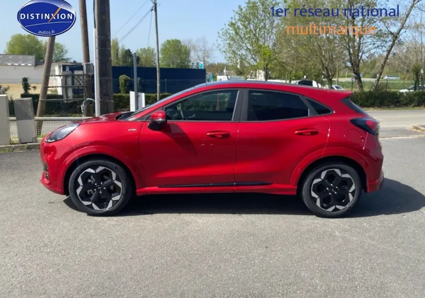 Profil côté gauche d'un Ford Puma rouge métallisé avec jantes alu noires au design hexagonal, stationné en extérieur.