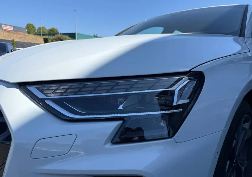 Gros plan sur le phare avant droit de l'Audi A3 Sportback blanc Glacier métallisé, avec jante alu visible en arrière-plan.