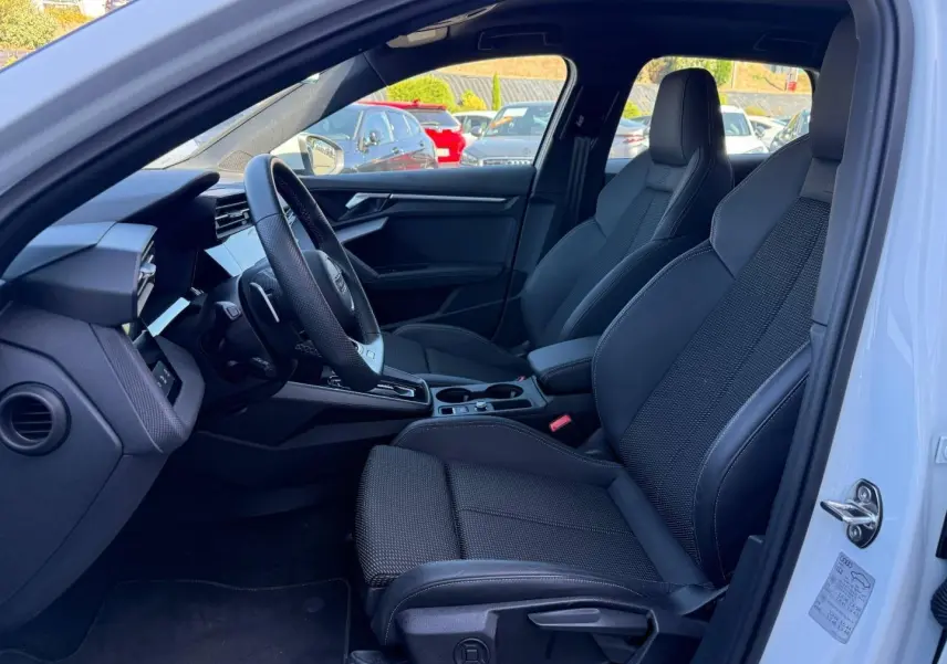 Vue intérieure côté conducteur de l'Audi A3 Sportback 2025 blanche, sièges sport noirs et volant multifonction cuir.