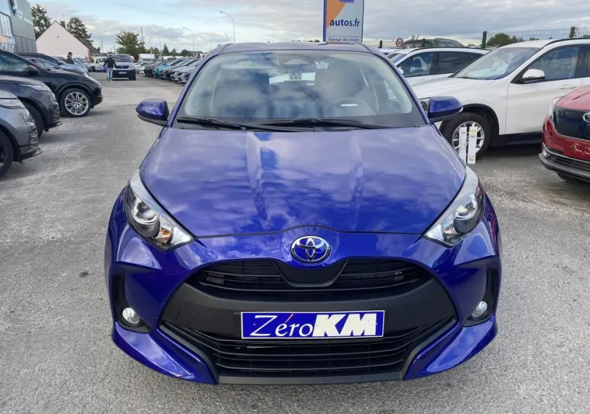 Vue frontale d'une Toyota Yaris 2025 bleu Genièvre avec calandre noire et phares LED allumés sur parking.