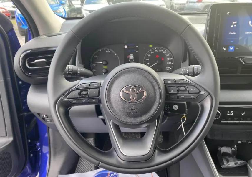 Vue centrée sur le volant noir de la Toyota Yaris 2025, tableau de bord et écran tactile avec portière bleu Genièvre ouverte.