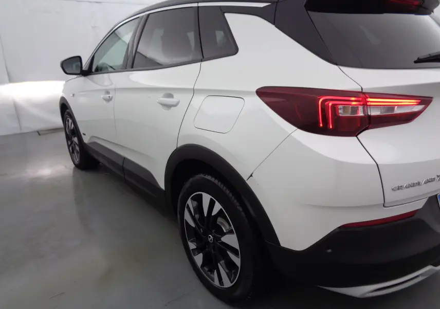 Vue 3/4 arrière côté gauche d'un Opel Grandland X blanc perle métallisé avec feux arrière allumés et jantes noires bicolores.