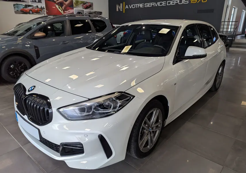 BMW Série 1 F40 18d blanc alpin vue 3/4 avant droit en showroom avec calandre noire et jantes alliage.