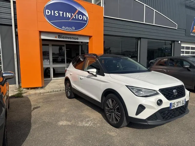 SEAT Arona 1.0 TSI Urban blanc Nevada avec toit gris Magnétique, vue 3/4 avant droit devant un garage Distinxion