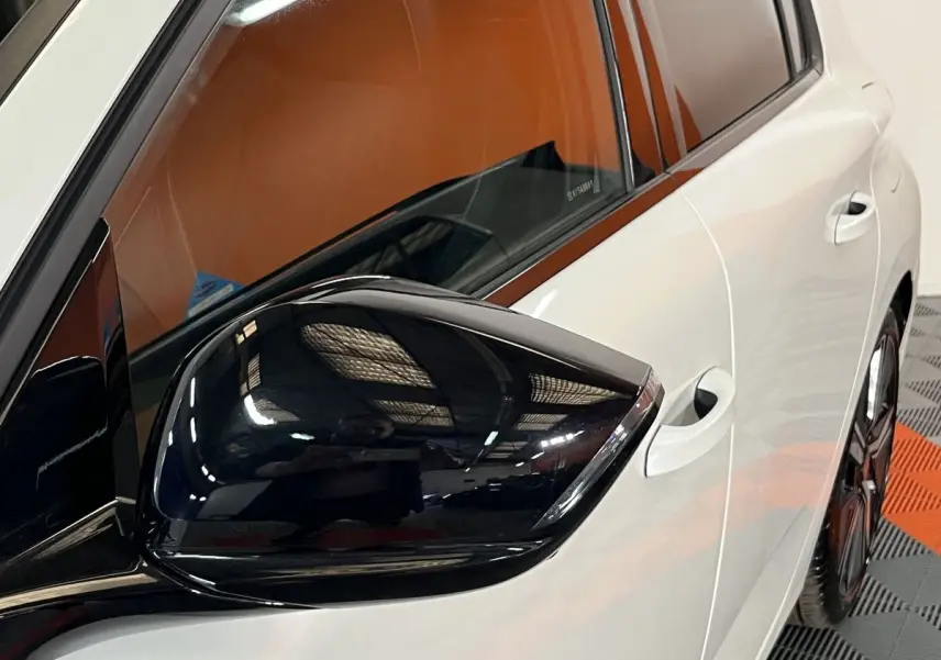 Gros plan sur le rétroviseur noir brillant côté gauche d'une Peugeot 308 GT PHEV blanc nacré, avec porte et vitre arrière visibles.
