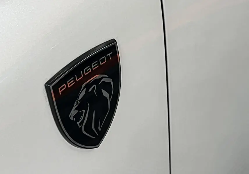 Gros plan sur l'écusson Peugeot noir et rouge apposé sur la carrosserie blanc nacré d'une 308 GT PHEV 2022.