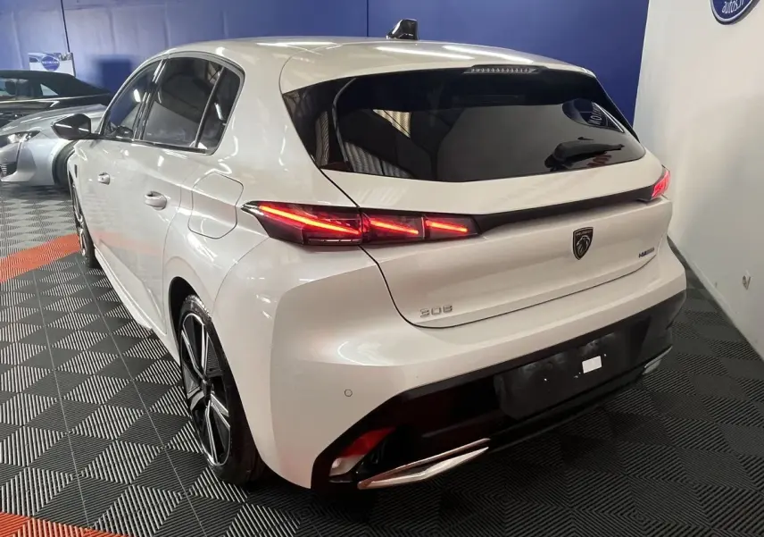 Vue 3/4 arrière droite d'une Peugeot 308 GT PHEV 180 blanche nacrée avec feux arrière LED allumés en intérieur showroom.
