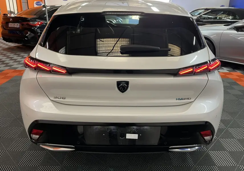 Vue arrière d'une Peugeot 308 GT PHEV blanche nacrée avec feux LED allumés et logo noir au centre du hayon.