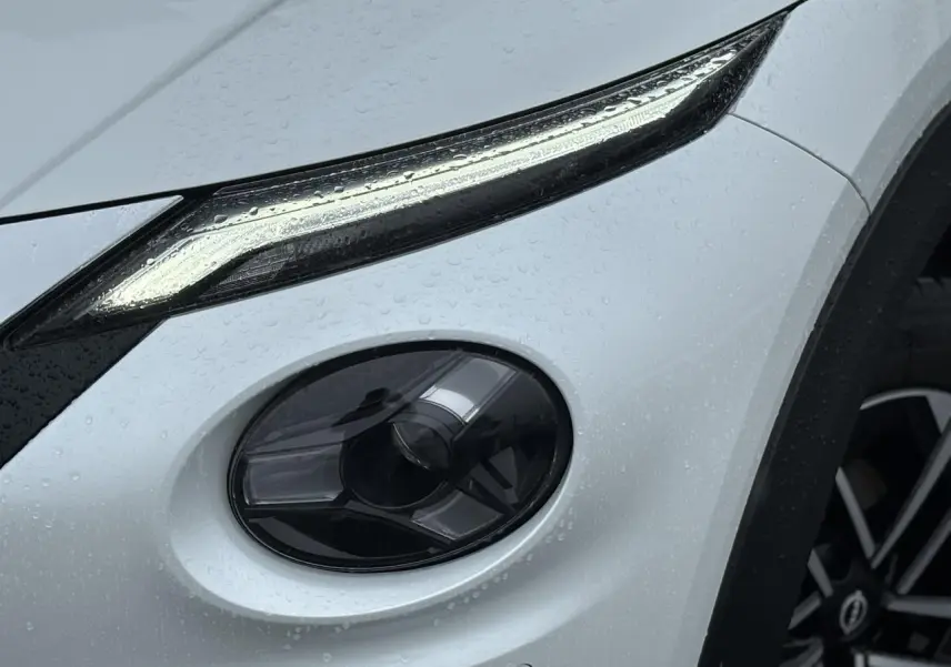 Gros plan sur le phare avant droit et la jante alliage 17'' d'un Nissan Juke blanc nacré sous la pluie.