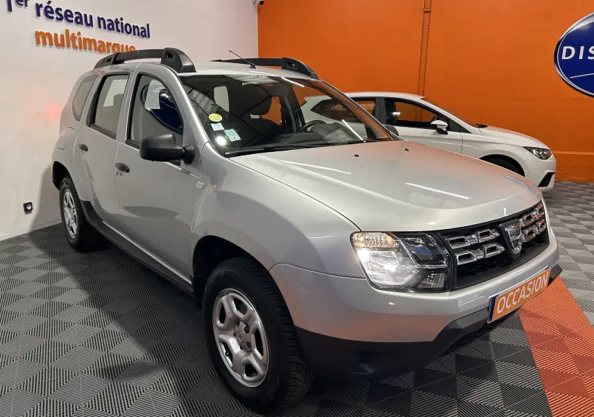 Vue 3/4 avant droit d'un Dacia Duster gris 2017 avec calandre chromée et barres de toit noires en showroom.