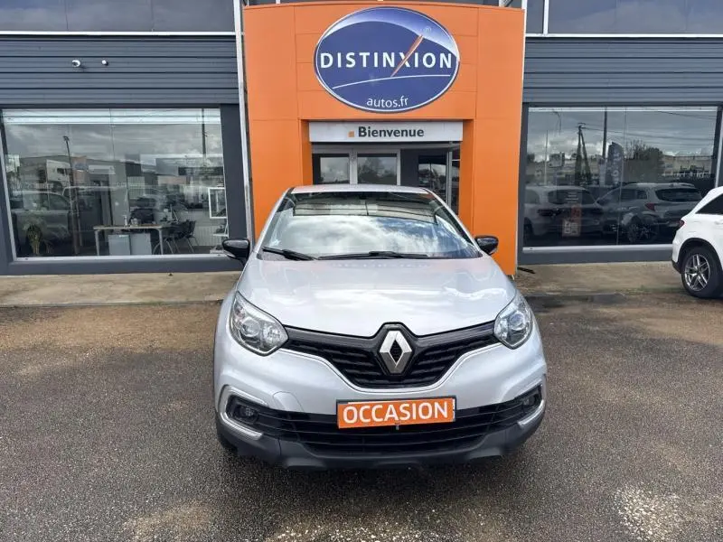 Vue avant d'un Renault Captur gris platine 2019 avec calandre noire et plaque orange "OCCASION" devant un garage Distinxion.