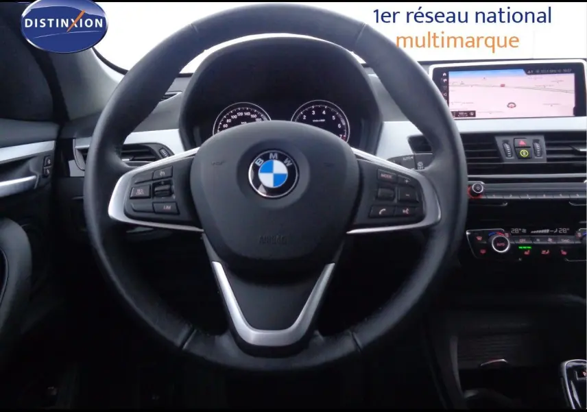 Vue centrée sur le volant BMW d'un X1 blanc Alpine 2022, avec tableau de bord numérique et écran tactile de navigation.