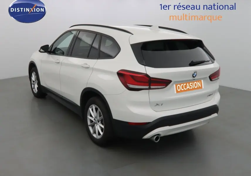 BMW X1 blanc Alpine métal vu en 3/4 arrière côté gauche dans un studio avec plaque occasion orange.