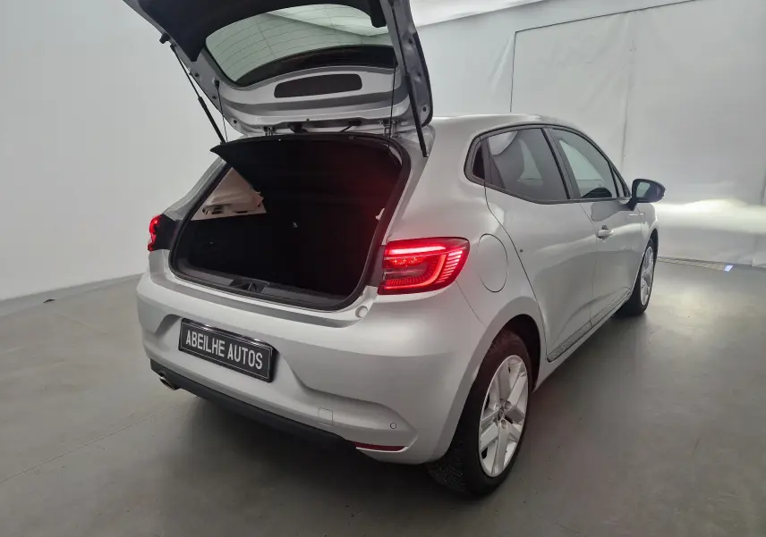 Vue 3/4 arrière droite d'une Renault Clio V gris platine avec coffre ouvert et feux arrière allumés dans un studio.