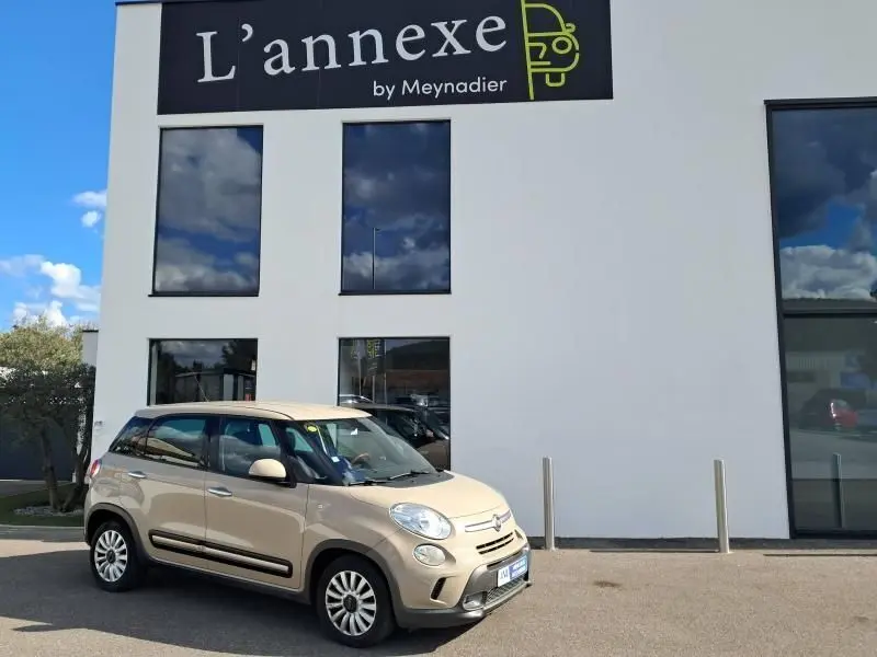 Vue 3/4 avant gauche d'une FIAT 500L beige 2014 stationnée devant un bâtiment blanc avec grandes fenêtres.