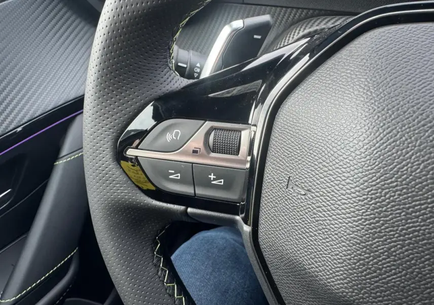 Gros plan sur le côté gauche du volant perforé du Peugeot 208 Hybrid 110 gris, avec commandes audio et finition noire brillante.