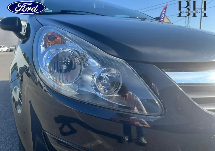 Gros plan sur le phare avant gauche d’une Opel Corsa noire de 2010, mettant en valeur la carrosserie brillante.
