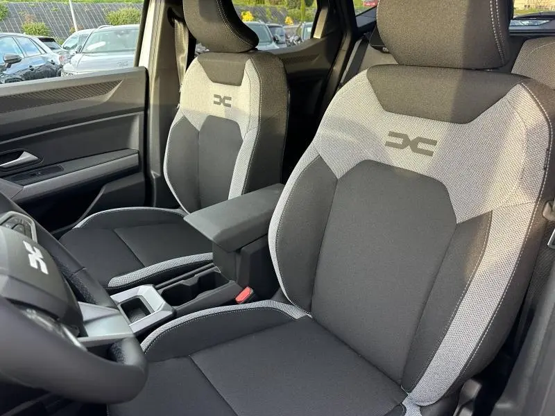 Vue intérieure du Dacia Duster 2025, sièges avant en tissu noir et gris avec logo DC, accoudoir central visible.