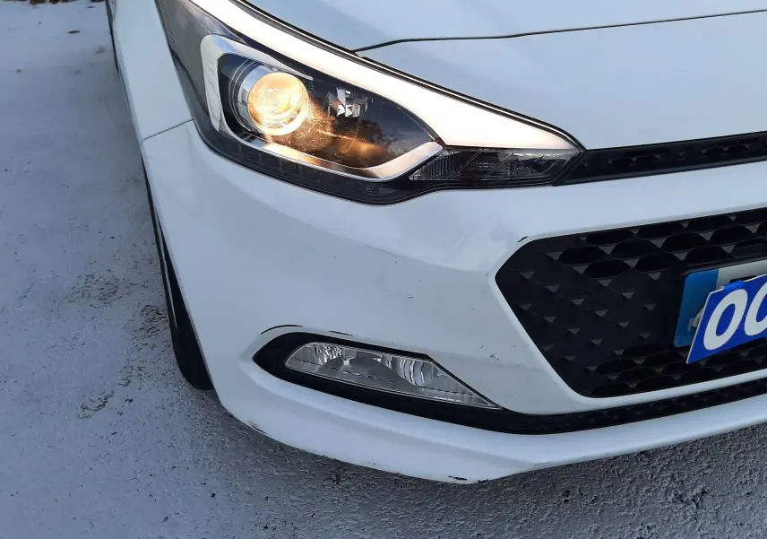 Gros plan sur l'avant droit blanc d'une Hyundai i20 2016 avec phare allumé et calandre noire ajourée.