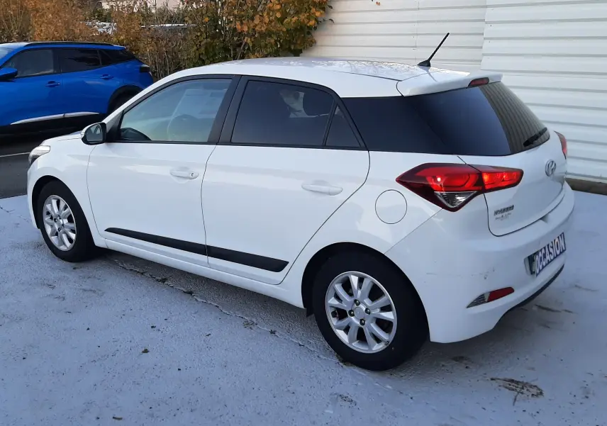 Hyundai i20 blanche vue en profil côté gauche, avec vitres arrière teintées et feux arrière allumés.