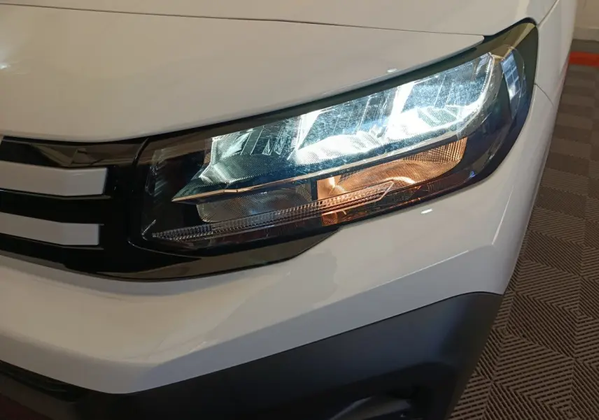 Gros plan sur le phare avant droit du Peugeot Partner blanc 2025, avec détails du contour noir et clignotant orange.