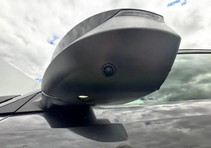 Gros plan sur le rétroviseur noir du côté gauche du Peugeot 5008 gris titane avec caméra intégrée sous un ciel nuageux.