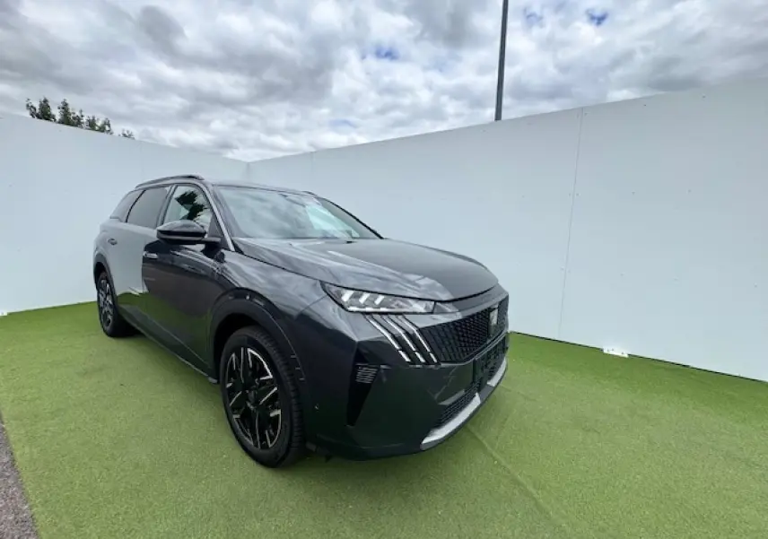 Peugeot 5008 gris titane toit noir en 3/4 avant droit sur fond vert et ciel nuageux.