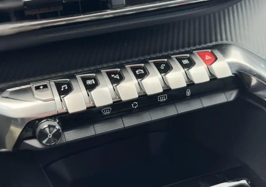 Gros plan sur la rangée de commandes argentées du tableau de bord du Peugeot 3008 noir, boutons fonctionnels et alerte rouge.
