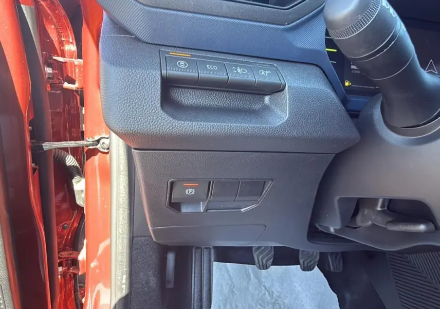 Vue intérieure côté gauche du tableau de bord du Dacia Duster orange 2025 avec commandes d'assistance et pédales visibles.