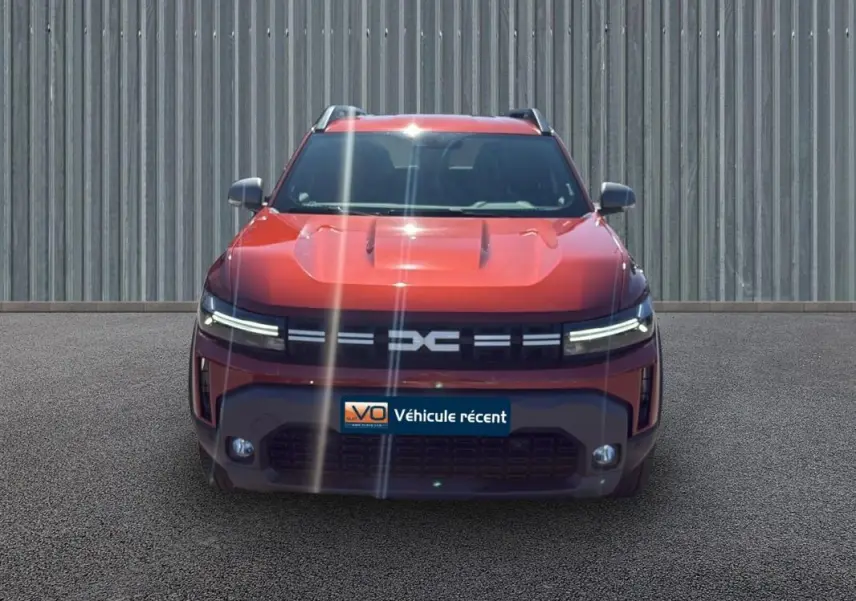 Vue frontale du Dacia Duster 2025 orange avec phares LED fins et barres de toit visibles sur fond urbain.