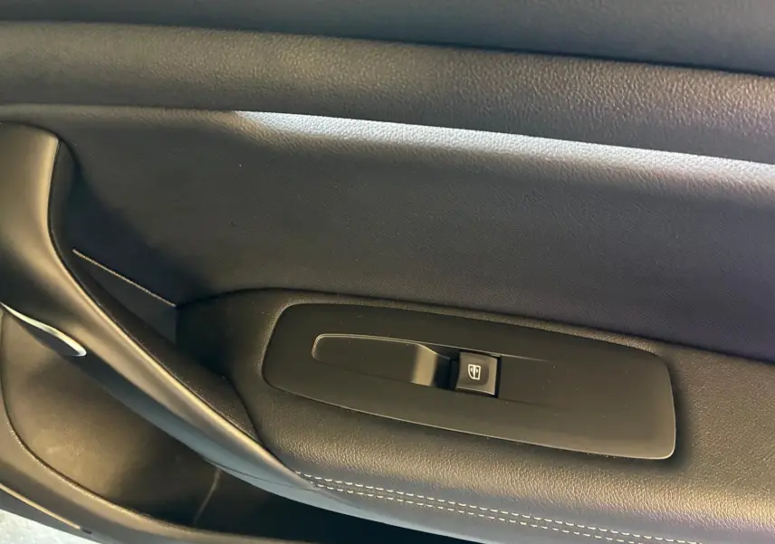 Gros plan sur la commande de lève-vitre côté gauche en plastique noir avec surpiqûres blanches sur la garniture intérieure.