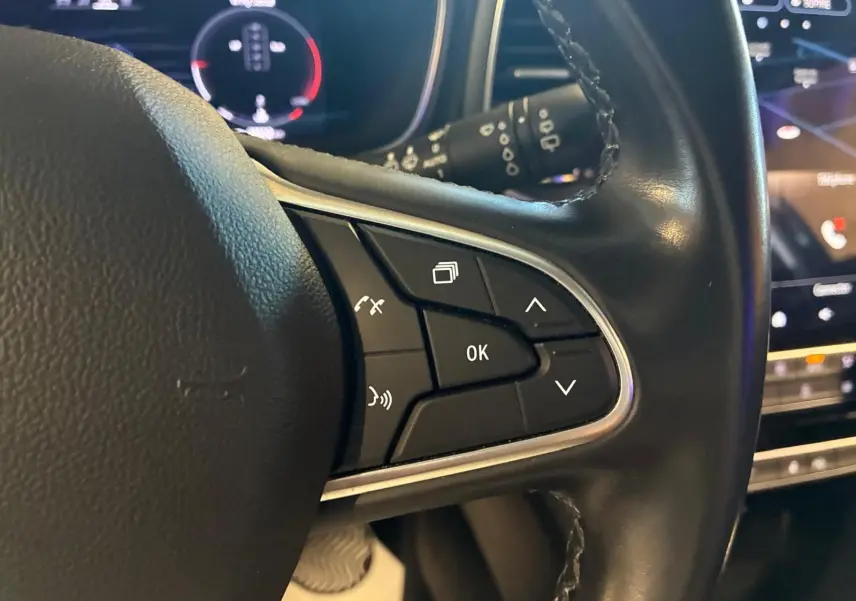 Gros plan sur les commandes au volant en cuir noir de la Renault Mégane 1.5 DCI 115 Intens, tableau de bord numérique en arrière-plan.