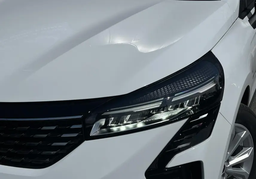 Gros plan sur le phare avant droit et la calandre noire d'une Renault Clio blanche Glacier avec jante 16 pouces.