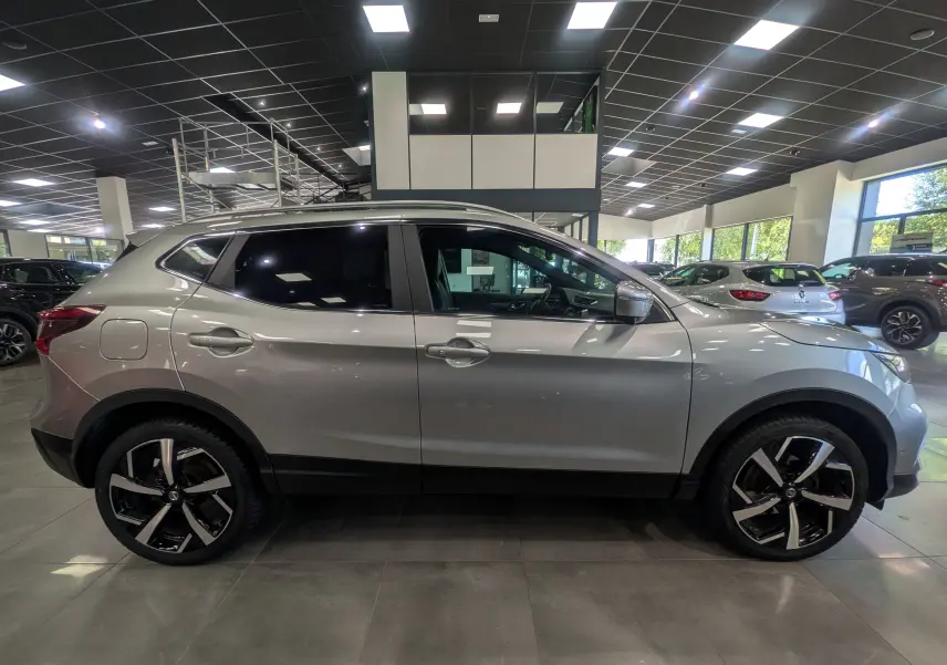 Vue latérale droite d'un Nissan Qashqai gris perle en showroom, avec jantes alliage bicolores et vitres teintées.