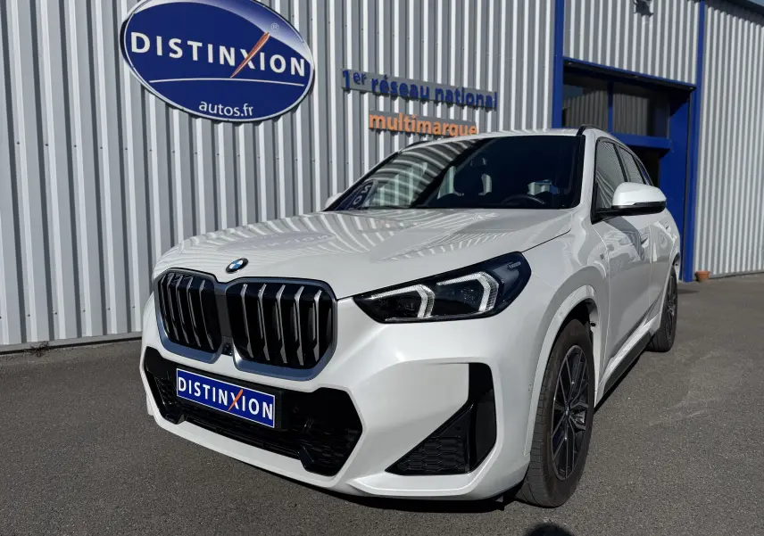 BMW X1 blanc minéral en 3/4 avant droit, avec calandre noire et phares LED distinctifs, stationné devant un bâtiment.