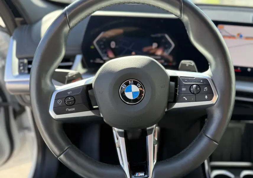 Gros plan sur le volant cuir noir du BMW X1 20D 2025 avec commandes multifonctions et tableau de bord numérique flou.