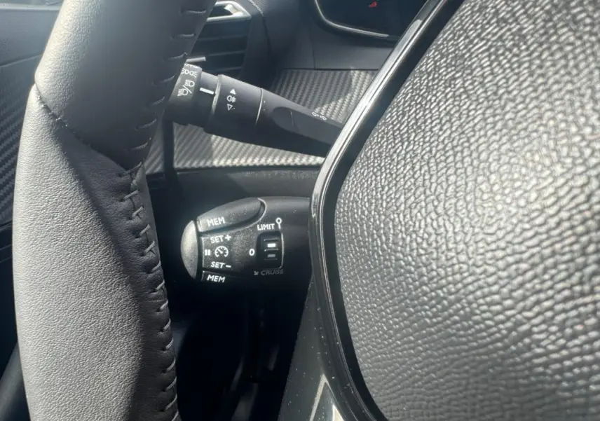 Gros plan sur la commande de régulateur de vitesse à gauche du volant dans l’habitacle noir du Peugeot 2008 blanc 2024.